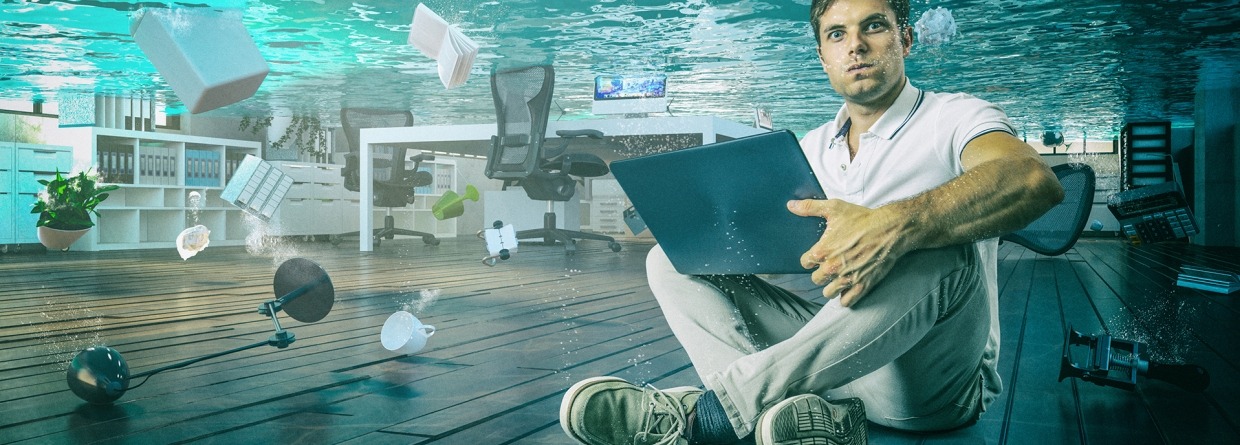 Man zit onder water gehurkt op een vloer met een laptop op schoot en zie je een lamp, bureau, plant, ordner en boek drijven