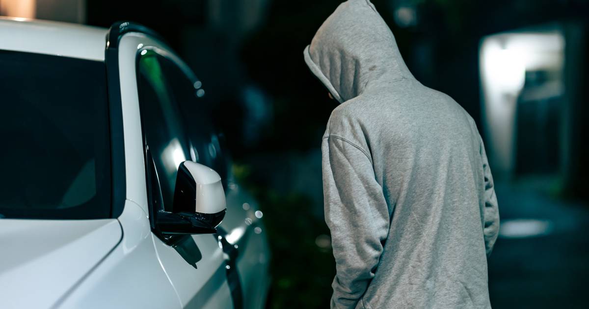 auto-inbraken; man met grijze hoodie aan en capuchon op loopt in het donker langs een witte auto en kijkt naar binnen 